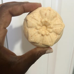 Peut inclure: Une bombe de bain ronde beige avec un motif floral. La bombe de bain a une surface textur&eacute;e et est parsem&eacute;e d'orange.