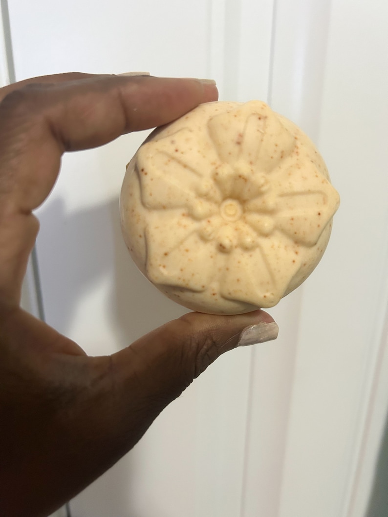 Peut inclure: Une bombe de bain ronde beige avec un motif floral. La bombe de bain a une surface textur&eacute;e et est parsem&eacute;e d'orange.