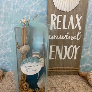 May include: A clear glass jar with a blue liquid and a silver lid. The jar is filled with brown paper shreds and has a small silver turtle charm attached to the top. The jar is sitting on a sandy surface with a wooden sign that says "Relax unwind enjoy" and a seashell.
