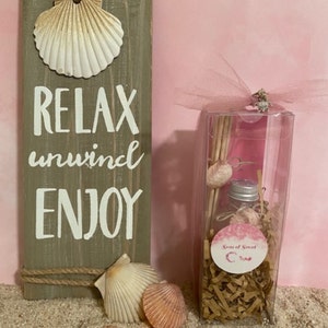May include: A clear glass bottle with a silver lid and a white label that says "Seas of Scent" is in a clear plastic box with brown shredded paper. The box is tied with a pink ribbon and a silver charm. The box is sitting on a sandy surface with seashells.