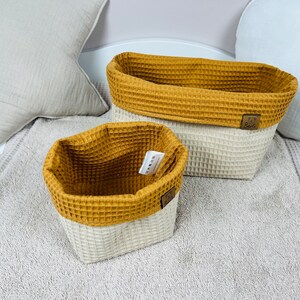 May include: Two woven storage baskets with a white base and a mustard yellow top. The baskets are made of a waffle-like fabric. The larger basket is rectangular and the smaller basket is square.