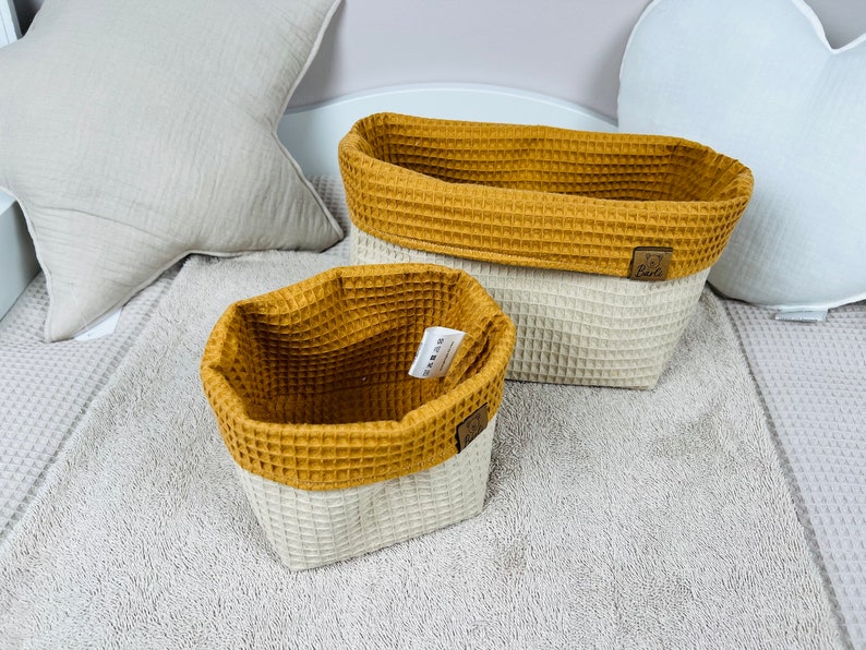May include: Two woven storage baskets with a white base and a mustard yellow top. The baskets are made of a waffle-like fabric. The larger basket is rectangular and the smaller basket is square.