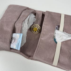 May include: A dusty rose corduroy nappy clutch featuring a dedicated pocket for wipes, a central compartment for nappies, and a cream-coloured elastic strap to secure the folded case. A beige dummy with a yellow teat is tucked inside.