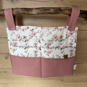 Könnte beinhalten: Ein rosa-weißer Stoff-Hängeregal mit vier Taschen. Der Organizer hat einen Blumendruck und hängt an einem Ast.