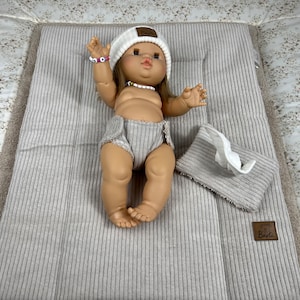 Puede incluir: Una muñeca de pelo castaño lleva un gorro de punto blanco con un parche Barli, una pulsera de cuentas y un cubrepañal de canalé beige. La muñeca está tumbada sobre un cambiador rectangular de canalé beige con un paño a juego y una etiqueta Barli.