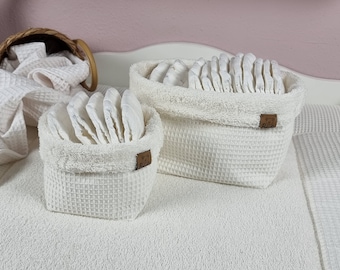 May include: Two off-white waffle-textured fabric baskets filled with folded white cloth diapers. Each basket features a small brown rectangular tag on the side. The baskets are placed on a matching white waffle-textured surface with extra fabric.
