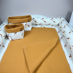 Puede incluir: Una cambiador blanco con estampado de jirafa marrón y blanco. El cambiador tiene una colchoneta de cambio de tejido de waffle amarillo mostaza y dos cestas de almacenamiento de tejido de waffle a juego.