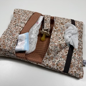 Pode incluir: Uma bolsa retangular de tecido para fraldas com um padrão floral em tons de castanho, bege e verde. O interior é forrado com tecido de textura waffle castanho e contém uma chupeta, uma fralda e um pacote de toalhitas com dispensador.
