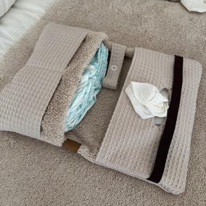 Peut inclure: Un tapis à langer en tissu beige à texture gaufrée doté d'une doublure intérieure douce, d'une sangle de fermeture à bouton-pression et d'une poche contenant une lingette blanche. Le tapis est rectangulaire avec une bande élastique marron.