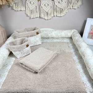 Könnte beinhalten: Eine beige Wickelunterlage mit Blumenmuster und einem gepolsterten beigefarbenen Rand. Zwei beigefarbene Aufbewahrungskörbe und ein beiges Handtuch liegen auf der Unterlage.
