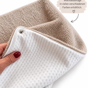 Op de afbeelding: Een beige, gestructureerde stoffen hoes met een wit wafelstof opzetstuk dat is bevestigd met een metalen drukknoop. De afbeelding bevat de tekst: Wechselauflage in vielen verschiedenen Farben erhältlich. met een hartje en een pijl.