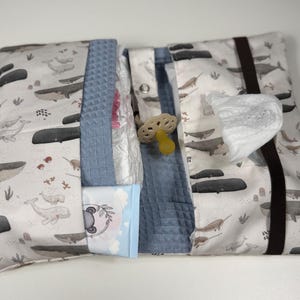 Puede incluir: Un estuche de tela plegable para pañales con un estampado de ballenas grises y negras sobre fondo blanco. El interior incluye bolsillos de tela azul con textura de gofre que contienen un pañal, un paquete de toallitas y un chupete de silicona beige.