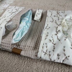 May include: A beige and white diaper bag with a floral pattern. The bag has three compartments, one for a diaper, one for wipes, and one for a small bottle of hand sanitizer.