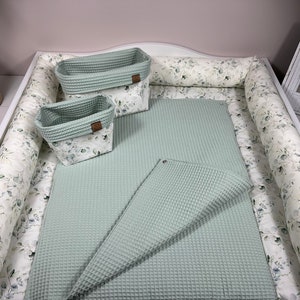Op de afbeelding: Een groene waffle weave verschoningsmat met twee witte en groene bloemenprint opbergmanden en een witte bloemenprint bumper.