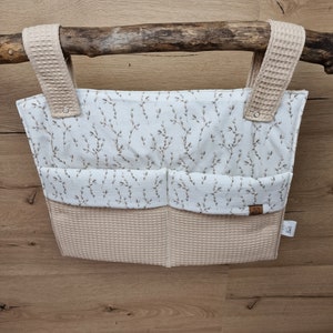 Può includere: Un organizzatore appeso beige e bianco con tre tasche. L'organizzatore ha un motivo floreale ed è realizzato in tessuto a nido d'ape. L'organizzatore è appeso a un ramo.