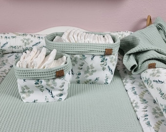 May include: Two fabric storage baskets with a white base featuring a green eucalyptus leaf pattern and a sage green waffle-textured rim. Each basket has a small brown rectangular tag attached to the side. The baskets are filled with white folded cloths.