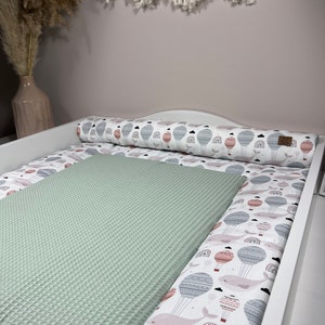 Könnte beinhalten: Eine weiße Wickelauflage mit einer grünen, wabenförmigen Wickelauflage darauf. Die Wickelauflage hat eine weiße Stütze mit einem Muster aus Heißluftballons und Walen.