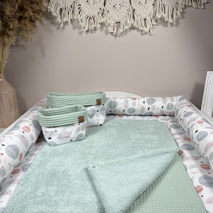 Può includere: Un fasciatoio bianco con un asciugamano verde chiaro e un asciugamano a nido d'ape. Il fasciatoio ha una stampa bianca e grigia di balene e palloncini. Ci sono due cesti di stoccaggio verde chiaro con una stampa di balene e palloncini.