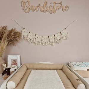 Könnte beinhalten: Eine beige Wickelauflage mit einer weißen Wickelunterlage und einem weißen und beigen Wickelkorb. Die Wickelauflage befindet sich auf einem weißen Wickeltisch. An der Wand hinter dem Wickeltisch befindet sich ein Holzschild mit der Aufschrift "Baerlistore". Über dem Wickeltisch hängt eine Makramee-Girlande. Auf der linken Seite des Bildes steht eine Vase mit getrocknetem Pampasgras. Auf dem Wickeltisch steht ein gerahmtes Foto eines Babys.