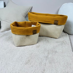May include: Two waffle-textured storage baskets, one rectangular and one oval, with a mustard yellow top and a cream bottom. The baskets are sitting on a fluffy white rug.