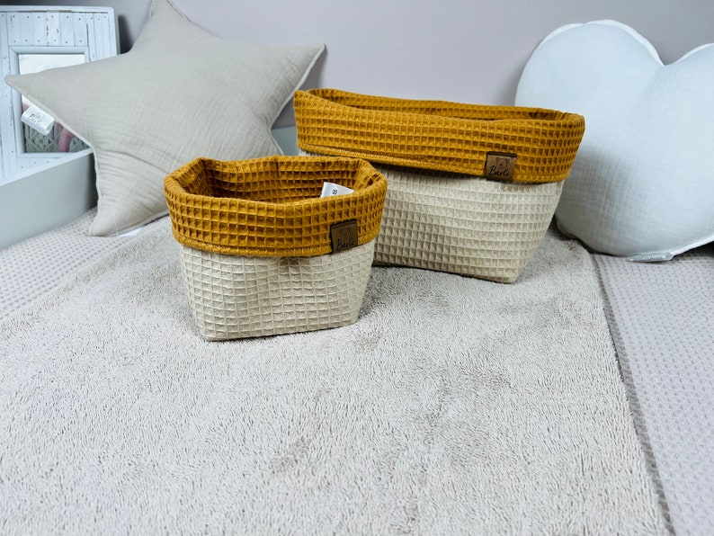 May include: Two waffle-textured storage baskets, one rectangular and one oval, with a mustard yellow top and a cream bottom. The baskets are sitting on a fluffy white rug.