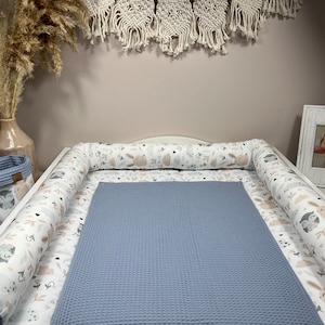 May include: A white changing pad with a blue waffle-textured changing mat. The changing pad has a white and beige print with small animal illustrations. The changing pad is surrounded by white and beige bolsters with the same animal print.