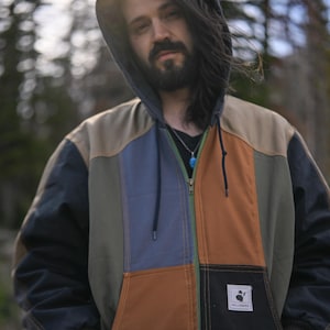 May include: A color-blocked zip-up hooded jacket featuring panels of tan, slate blue, olive green, brown, and black fabric. The jacket includes a front zipper, ribbed cuffs, and a white rectangular patch on the lower pocket with the text HELLEBORE.