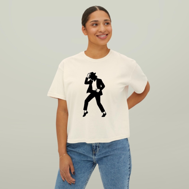 Puede incluir: Camiseta color crema con una silueta negra de una persona con traje y sombrero, en pose de baile. El gr&aacute;fico est&aacute; centrado en la camiseta. La modelo lleva vaqueros azules.