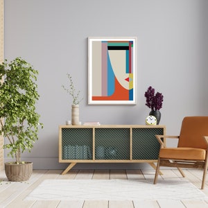 May include: A living room interior with a light grey wall, a wooden cabinet with a wire mesh front, a brown armchair, a potted plant, and a framed abstract geometric print with a red, blue, and yellow color scheme.