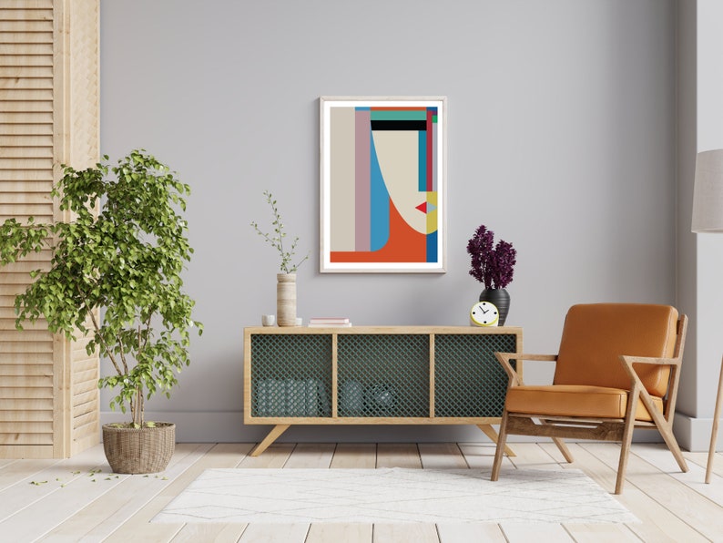 May include: A living room interior with a light grey wall, a wooden cabinet with a wire mesh front, a brown armchair, a potted plant, and a framed abstract geometric print with a red, blue, and yellow color scheme.