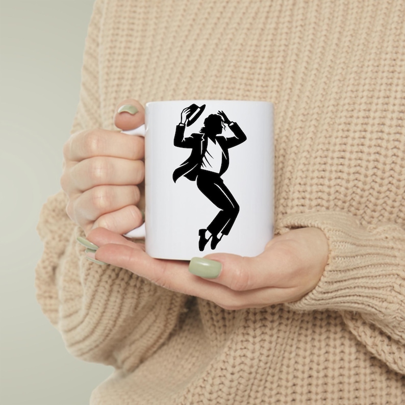 Puede incluir: Taza de cer&aacute;mica blanca con una silueta negra de una persona bailando, sosteniendo un sombrero. La persona lleva un traje. La taza tiene un asa est&aacute;ndar y la sostiene una persona que lleva un su&eacute;ter beige.