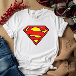May include: White t-shirt featuring the Superman logo. The logo is a red diamond with a yellow 'S' in the center, outlined in black. The shirt is made of a soft material and is suitable for casual wear.