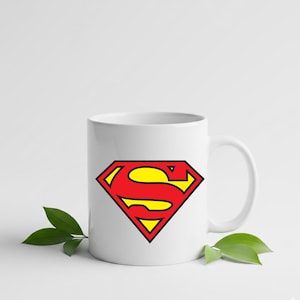 May include: White ceramic mug featuring the iconic Superman logo. The logo is a red diamond shape with a yellow 'S' inside, outlined in black. The mug has a curved handle and is suitable for hot or cold beverages.