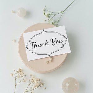 May include: A white card with black lettering that says "Thank You" in a decorative font. The card is sitting on a light pink round surface. There are small white flowers and two white round stones on a white background.