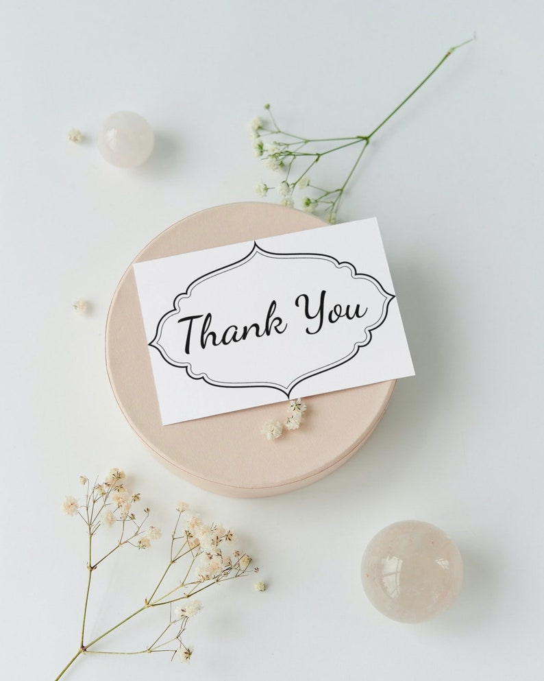 May include: A white card with black lettering that says "Thank You" in a decorative font. The card is sitting on a light pink round surface. There are small white flowers and two white round stones on a white background.