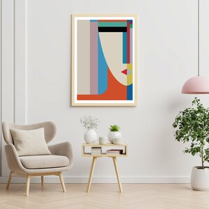 May include: Abstract geometric art print with a stylized face in shades of blue, red, yellow, and green. The print is framed in a light wood frame and is hanging on a white wall.