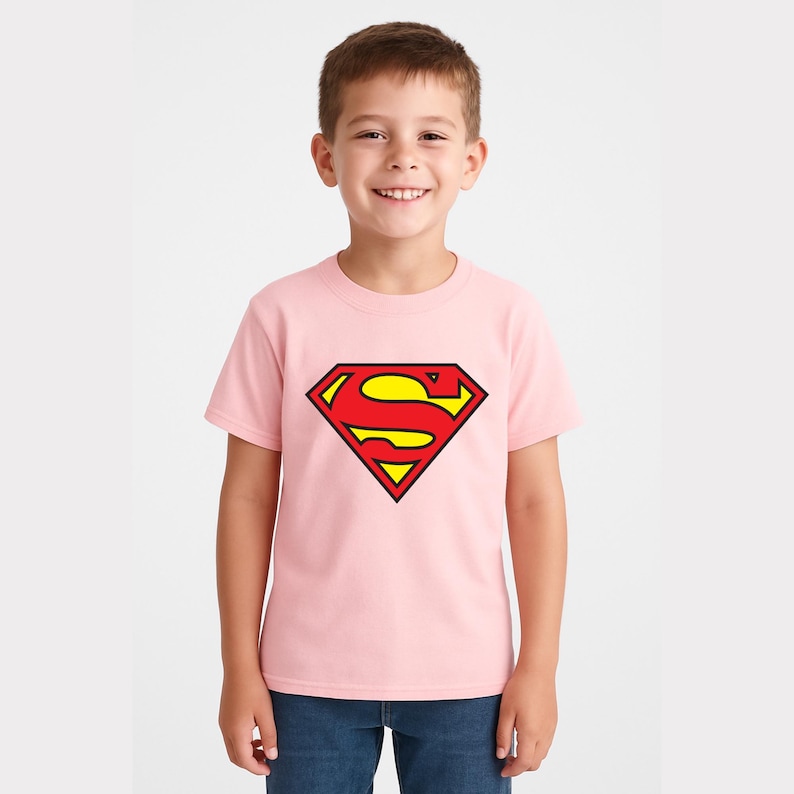May include: A light pink t-shirt featuring the Superman logo in red, yellow, and black. The iconic "S" shield is prominently displayed on the front of the shirt. The shirt is made of a soft material and is suitable for children.