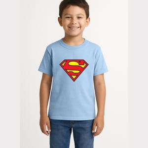 May include: A light blue t-shirt featuring the Superman logo in red and yellow. The logo is a diamond shape with the letter "S" in the center. The shirt is short-sleeved and made of a soft material.