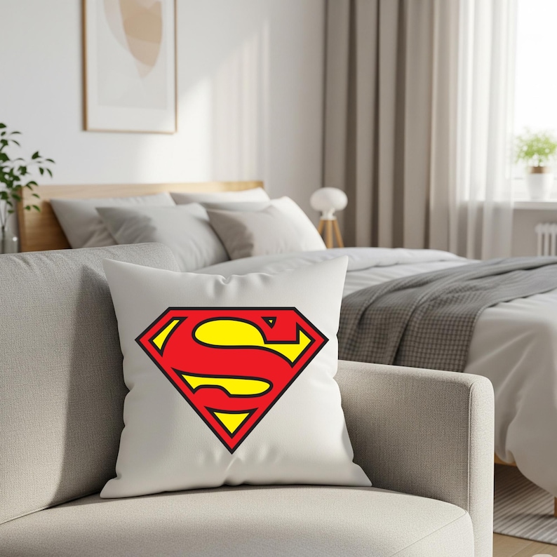 May include: White throw pillow featuring the iconic Superman logo. The logo is a red 'S' on a yellow diamond, outlined in black. The pillow is placed on a light gray sofa in a bedroom setting.