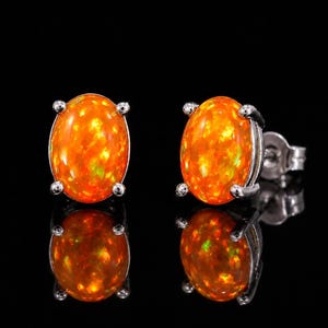 May include: A pair of oval-shaped stud earrings featuring vibrant orange fire opals set in silver-toned metal prongs. Each earring displays a smooth, polished cabochon surface with internal flashes of yellow and green light against a dark background.