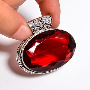 May include: Oval-shaped, faceted, deep red gemstone pendant with a decorative silver-colored bail. The pendant is held by a hand against a white background. The gemstone has a brilliant cut, reflecting light. The silver setting has intricate detailing.