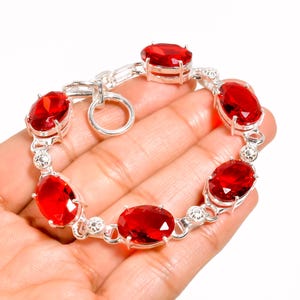 Könnte beinhalten: Ein silbernes Armband mit ovalen, leuchtend roten Edelsteinen. Das Armband hat ein kettenartiges Design, wobei die Edelsteine in einzelnen Silberfassungen gefasst sind. Der Verschluss ist ein einfacher Ring- und Hakenverschluss. Das Armband ist ein Schmuckstück.