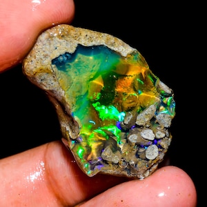 May include: A rough-cut opal gemstone with vibrant colors of green, yellow, and blue. The opal is held in a hand, showcasing its natural, unpolished texture and the play of light across its surface. The stone is surrounded by a matrix of brown and gray rock.