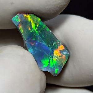 May include: A striking, uncut opal gemstone with a dark background. The opal exhibits a play of colour, with flashes of green, blue, yellow, and orange. The stone is held by a gloved hand, highlighting its natural, irregular form.