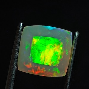 May include: A square-cut opal gemstone featuring a translucent body with a vibrant play-of-color, displaying intense flashes of neon green, yellow, and orange throughout its center, held by dark metal tweezers against a solid black background.