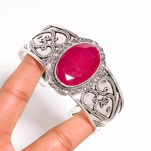 May include: A silver cuff bracelet with an oval, faceted, ruby-red gemstone. The bracelet has an ornate, filigree design with scroll and heart motifs. The gemstone is the focal point of the bracelet.