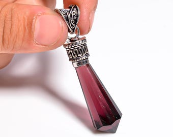Puede incluir: Un colgante facetado de color rojo intenso con una fianza plateada ornamentada. El colgante tiene una forma geométrica alargada, que se estrecha hasta un punto. La fianza presenta remolinos decorativos y un pequeño lazo para una cadena. El colgante se sostiene sobre un fondo blanco.