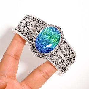 May include: Silver cuff bracelet with an oval, iridescent blue and green stone. The bracelet has an ornate, filigree design on the band and around the stone setting. The jewellery is held by a hand against a white background.
