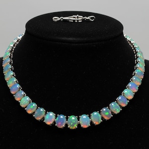 Puede incluir: Un collar de plata con una serie de ópalos ovalados. Los ópalos muestran un juego de colores con tonos azules, verdes y rojos. El collar se exhibe sobre un fondo negro, resaltando el brillo de las piedras preciosas.
