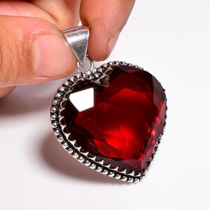 May include: A heart-shaped pendant featuring a large, faceted, deep red gemstone. The gemstone is set in a silver-coloured metal frame with a decorative beaded edge and a triangular bail for a chain.